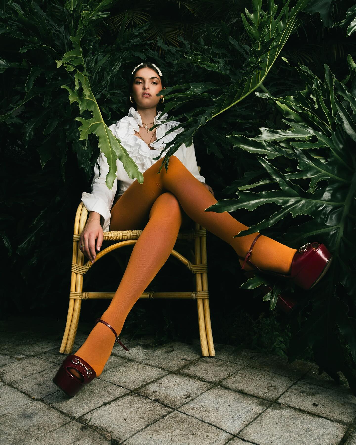 Editorial para InStyle Grecia: modelo sentada entre plantas tropicales, medias naranjas y tacones burdeos.