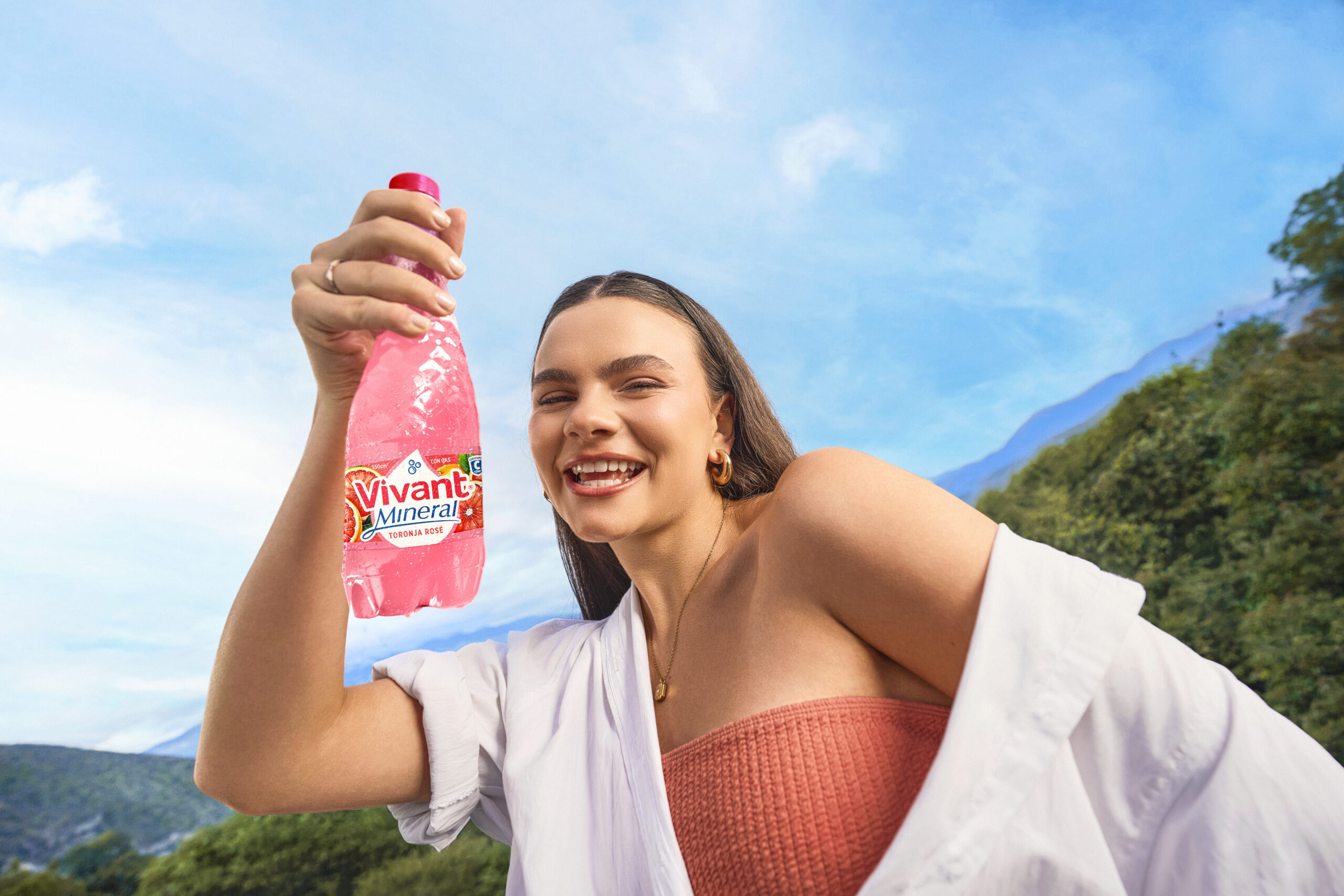 Modelo sonriente sosteniendo una botella de Vivant Mineral sabor toronja rosé frente a montañas.