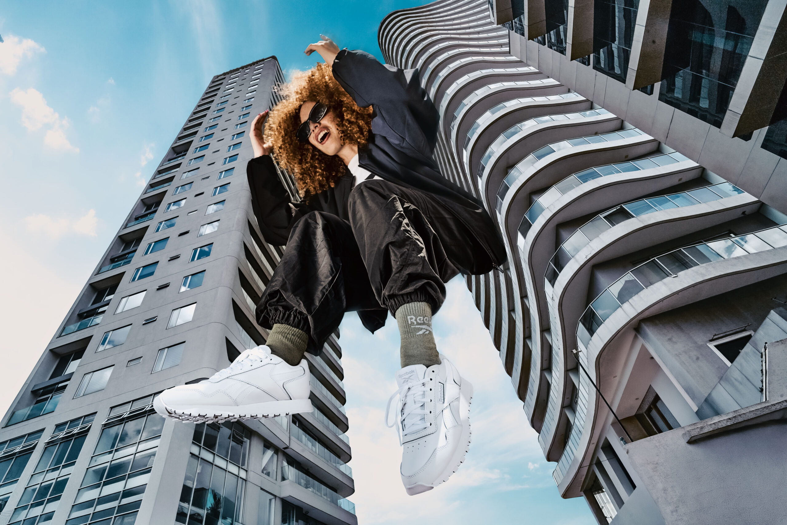 Modelo saltando con zapatillas Reebok blancas, fotografiada en contrapicado entre dos edificios.